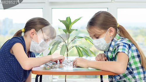 Image of Two girls in medical masks sit at a table and play board games.