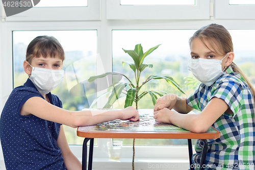 Image of Two girlfriends in medical masks play puzzles and looked into the frame.