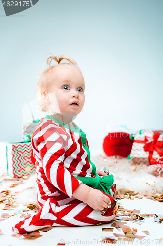 Image of Cute baby girl 1 year old near santa hat posing over Christmas background. Sitting on floor with Christmas ball. Holiday season.