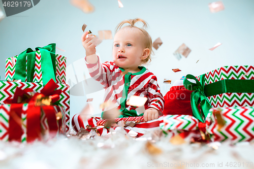 Image of Cute baby girl 1 year old near santa hat posing over Christmas background. Sitting on floor with Christmas ball. Holiday season.