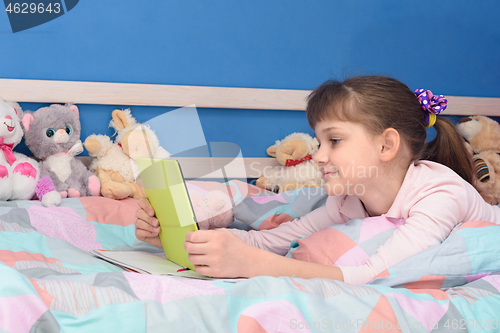Image of The girl lies in bed at home on her stomach and looks at the screen of the tablet