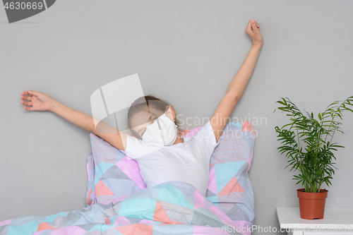 Image of A recovering girl in a medical mask woke up in the morning and stretches herself in bed