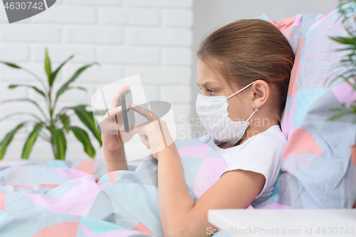 Image of The girl lies in bed in a medical mask and does web surfing on the phone