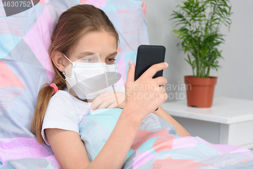 Image of A girl in a medical mask lies in bed and looks at the phone screen