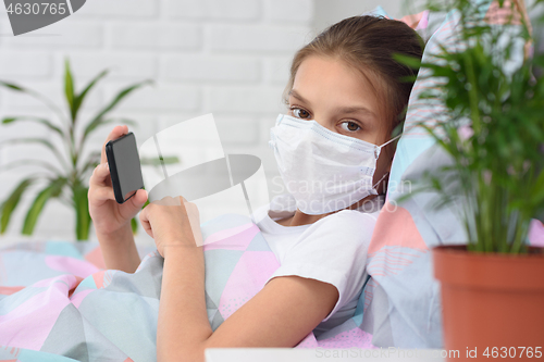 Image of The girl lies on the bed in a medical mask with a mobile phone and looked into the frame