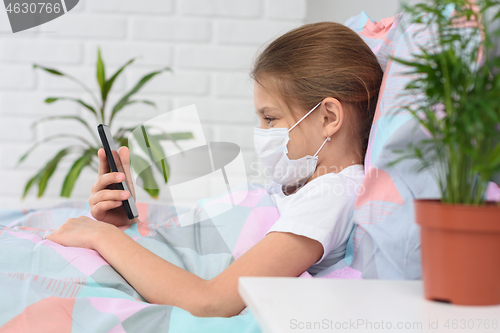 Image of A girl in a hospital ward lies on a bed in a medical mask and does web surfing on the phone
