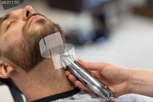 Image of man and barber with trimmer cutting beard at salon