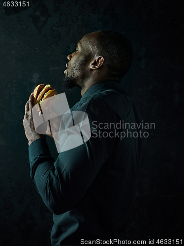 Image of young african american man eating hamburger and looking away on black studio