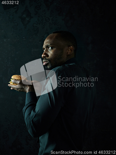 Image of young african american man eating hamburger and looking away on black studio