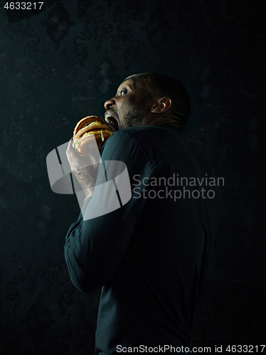 Image of young african american man eating hamburger and looking away on black studio