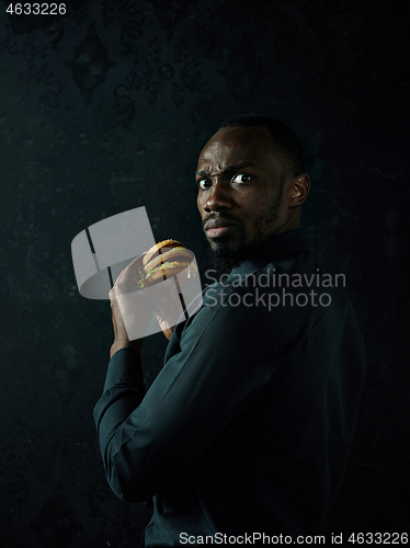 Image of young african american man eating hamburger and looking away on black studio
