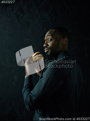 Image of young african american man eating hamburger and looking away on black studio