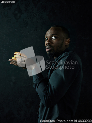 Image of young african american man eating hamburger and looking away on black studio