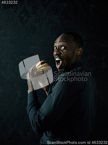 Image of young african american man eating hamburger and looking away on black studio