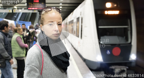 Image of Young woman in winter coat going to work, waiting on the platform of a railway station for train to arrive. Public transport