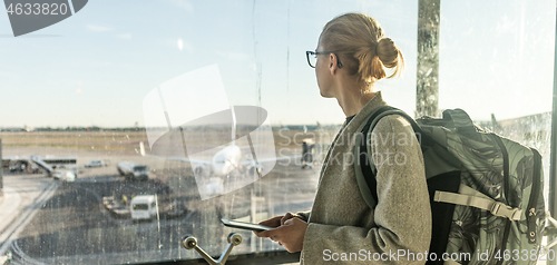 Image of Young woman standing near airport gates window holding cellphone in her hands, wearing travel backpack and walking to lounge area. Female traveler search online map at the airport