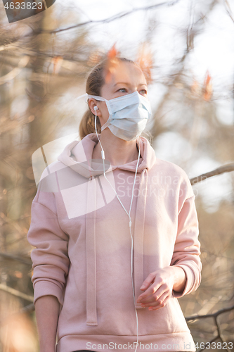 Image of Portrait of caucasian sporty woman wearing medical protection face mask while walking in park, relaxing and listening to music. Corona virus, or Covid-19, is spreading all over the world