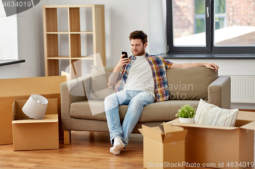 Image of man with smartphone and boxes on sofa at new home