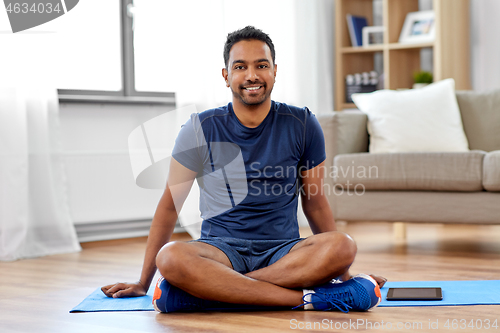 Image of smiling indian man sitting on exercise mat at home