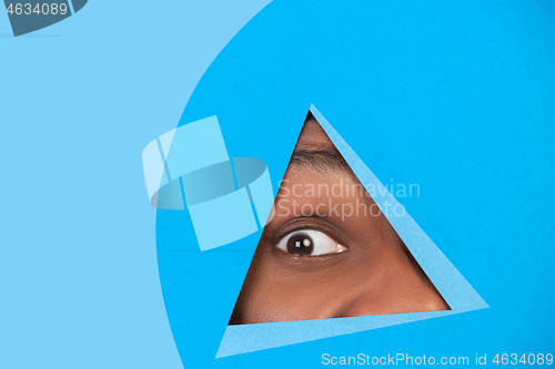 Image of Eye of african-american man peeking throught triangle in blue background