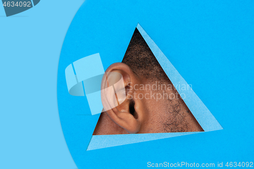 Image of Ear of african-american man peeking throught triangle in blue background