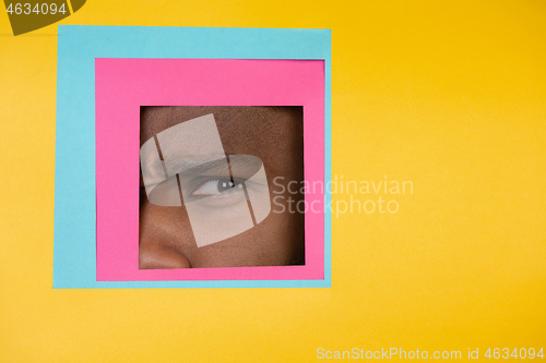 Image of Eye of african-american man peeking throught square in yellow background