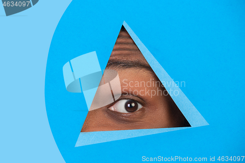 Image of Eye of african-american man peeking throught triangle in blue background