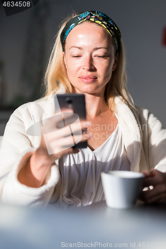 Image of Beautiful caucasian woman at home, feeling comfortable wearing white bathrobe, taking some time to herself, drinking morning coffee and reading news on mobile phone device in the morning