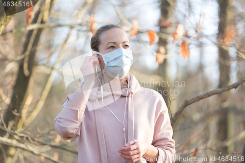 Image of Portrait of caucasian sporty woman wearing medical protection face mask while walking in park, relaxing and listening to music. Corona virus, or Covid-19, is spreading all over the world