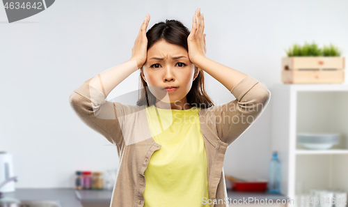 Image of stressed asian woman holding to head at kitchen