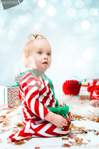 Image of Cute baby girl 1 year old near santa hat posing over Christmas background. Sitting on floor with Christmas ball. Holiday season.