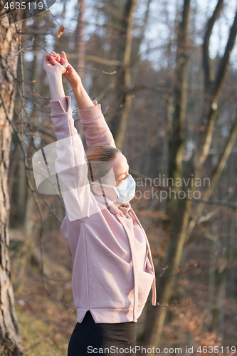 Image of Portrait of caucasian sporty woman wearing medical protection face mask while relaxing by stretching in forest. Corona virus, or Covid-19, is spreading all over the world