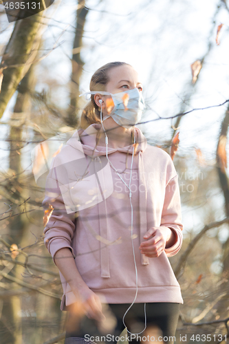 Image of Portrait of caucasian sporty woman wearing medical protection face mask while walking in park, relaxing and listening to music. Corona virus, or Covid-19, is spreading all over the world