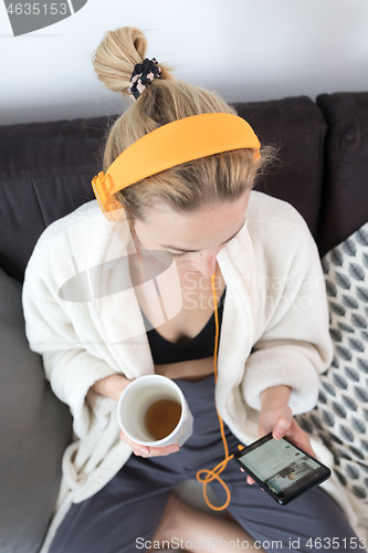 Image of Stay at home. Social distancing. Woman at home relaxing on sofa couch drinking tea from white cup, listening to relaxing music, stay connected to friens and family via social networks on mobile.