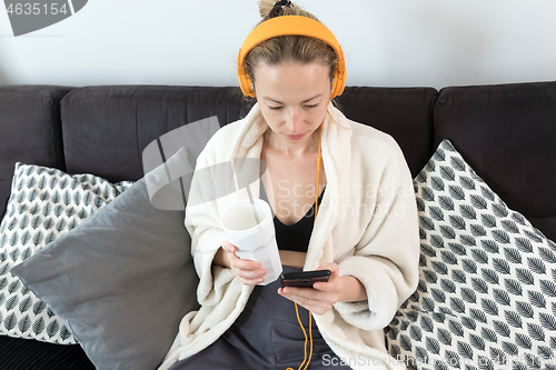 Image of Stay at home. Social distancing. Woman at home relaxing on sofa couch drinking tea from white cup, listening to relaxing music, stay connected to friens and family via social networks on mobile.