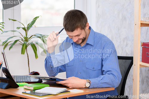 Image of Office employee reads a document, smiles and scratches his head with a pen