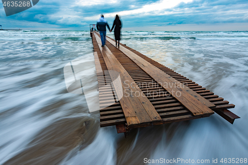 Image of Two people go down the wooden sea pier into the distance, evening, long exposure