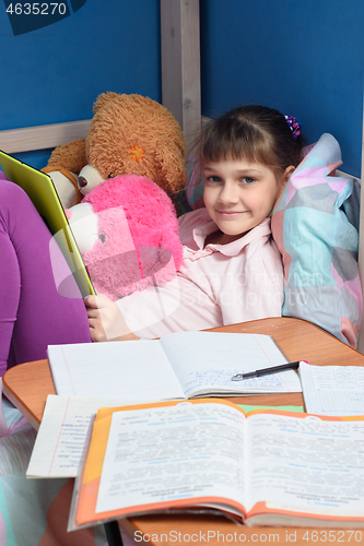 Image of The girl looks into the frame with a smile holding a tablet, in front of her lie textbooks and notebooks