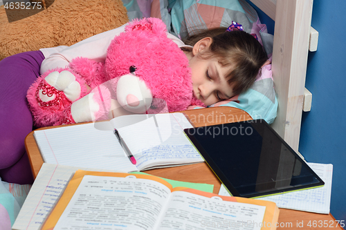 Image of Girl sleeps with a teddy bear near a desk with textbooks and a tablet