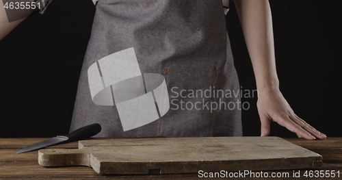 Image of Chef in a apron are standing at the table with wooden cutting board on it on a black background.