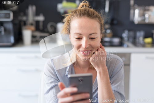Image of Young smiling cheerful pleased woman indoors at home kitchen using social media apps on mobile phone for chatting and stying connected with her loved ones. Stay at home, social distancing lifestyle.