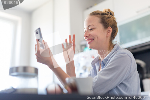 Image of Young smiling cheerful pleased woman indoors at home kitchen using social media on mobile phone for chatting and stying connected with her loved ones. Stay at home, social distancing lifestyle.