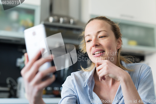 Image of Young smiling cheerful pleased woman indoors at home kitchen using social media apps on mobile phone for chatting and stying connected with her loved ones. Stay at home, social distancing lifestyle.