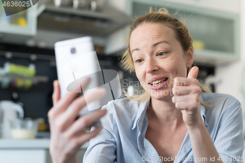 Image of Young smiling cheerful pleased woman indoors at home kitchen using social media apps on mobile phone for chatting and stying connected with her loved ones. Stay at home, social distancing lifestyle.