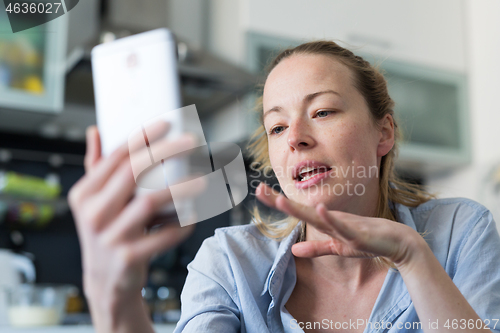 Image of Young smiling cheerful pleased woman indoors at home kitchen using social media apps on mobile phone for chatting and stying connected with her loved ones. Stay at home, social distancing lifestyle.