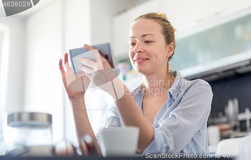 Image of Young smiling cheerful pleased woman indoors at home kitchen using social media on mobile phone for chatting and stying connected with her loved ones. Stay at home, social distancing lifestyle.