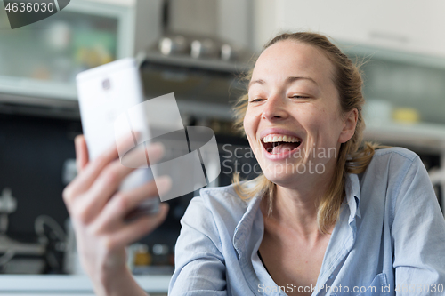 Image of Young smiling cheerful pleased woman indoors at home kitchen using social media apps on mobile phone for chatting and stying connected with her loved ones. Stay at home, social distancing lifestyle.