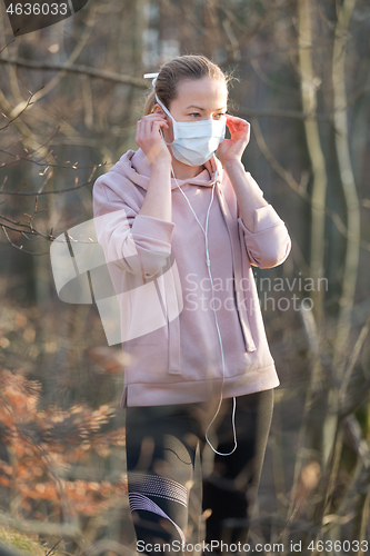 Image of Portrait of caucasian sporty woman wearing medical protection face mask while walking in park, relaxing and listening to music. Corona virus, or Covid-19, is spreading all over the world