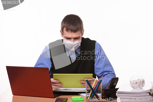 Image of Office worker in a medical mask at the workplace looks at the tablet screen