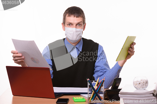 Image of A man in a medical mask holds a paper document in one hand, and a tablet computer in the other